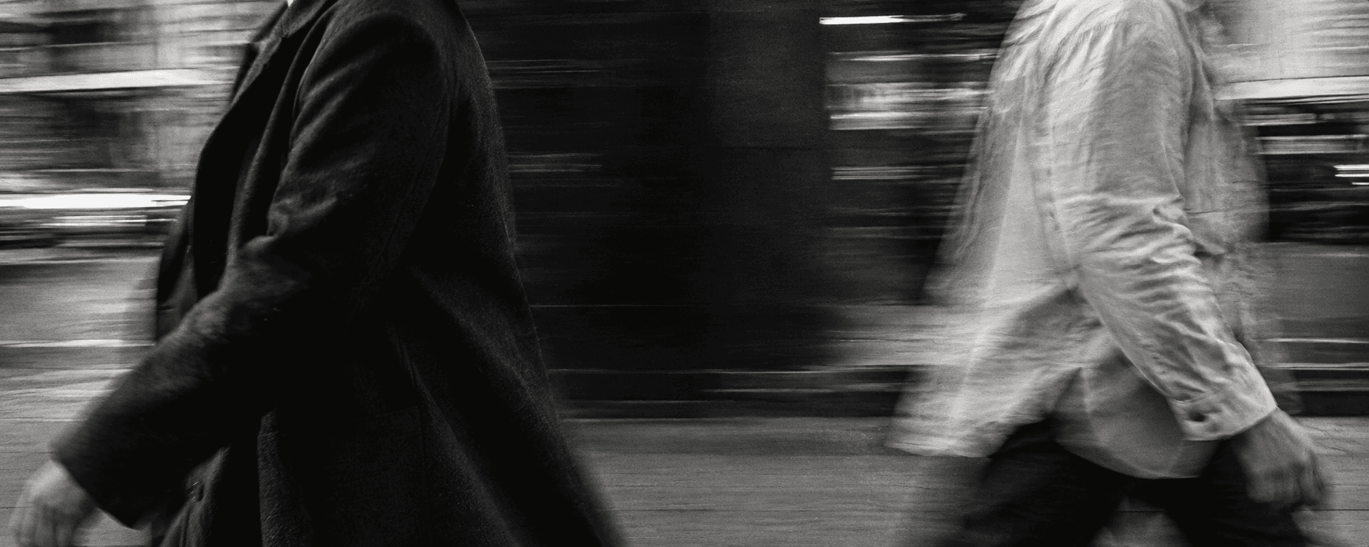 Black-and-white editorial image of two people passing, captured with film grain and motion blur